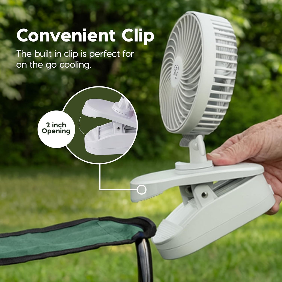 White clip-on fan with a hand holding it, set against a green outdoor background.