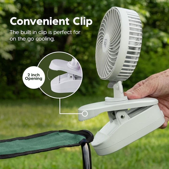 White clip-on fan with a hand holding it, set against a green outdoor background.