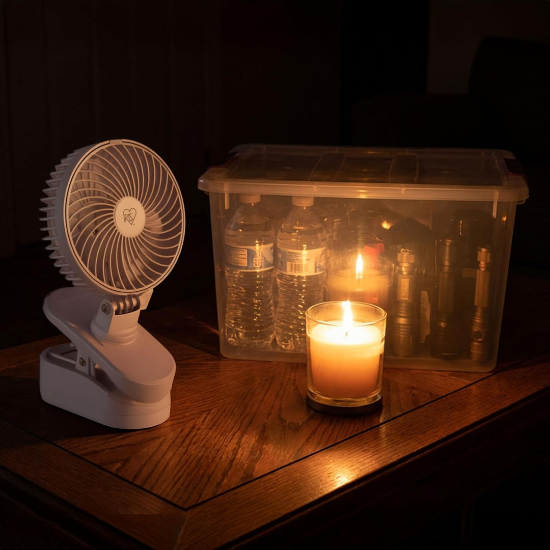 Small fan, plastic container with bottles, and lit candle on a wooden surface in low light.